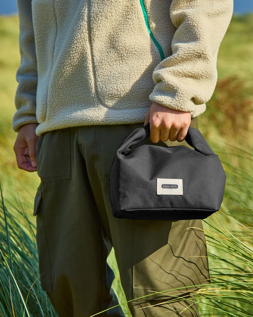 Maiden Cricket Black+Blum insulated lunch bag in black with roll-top closure held by a person in a grassy field
