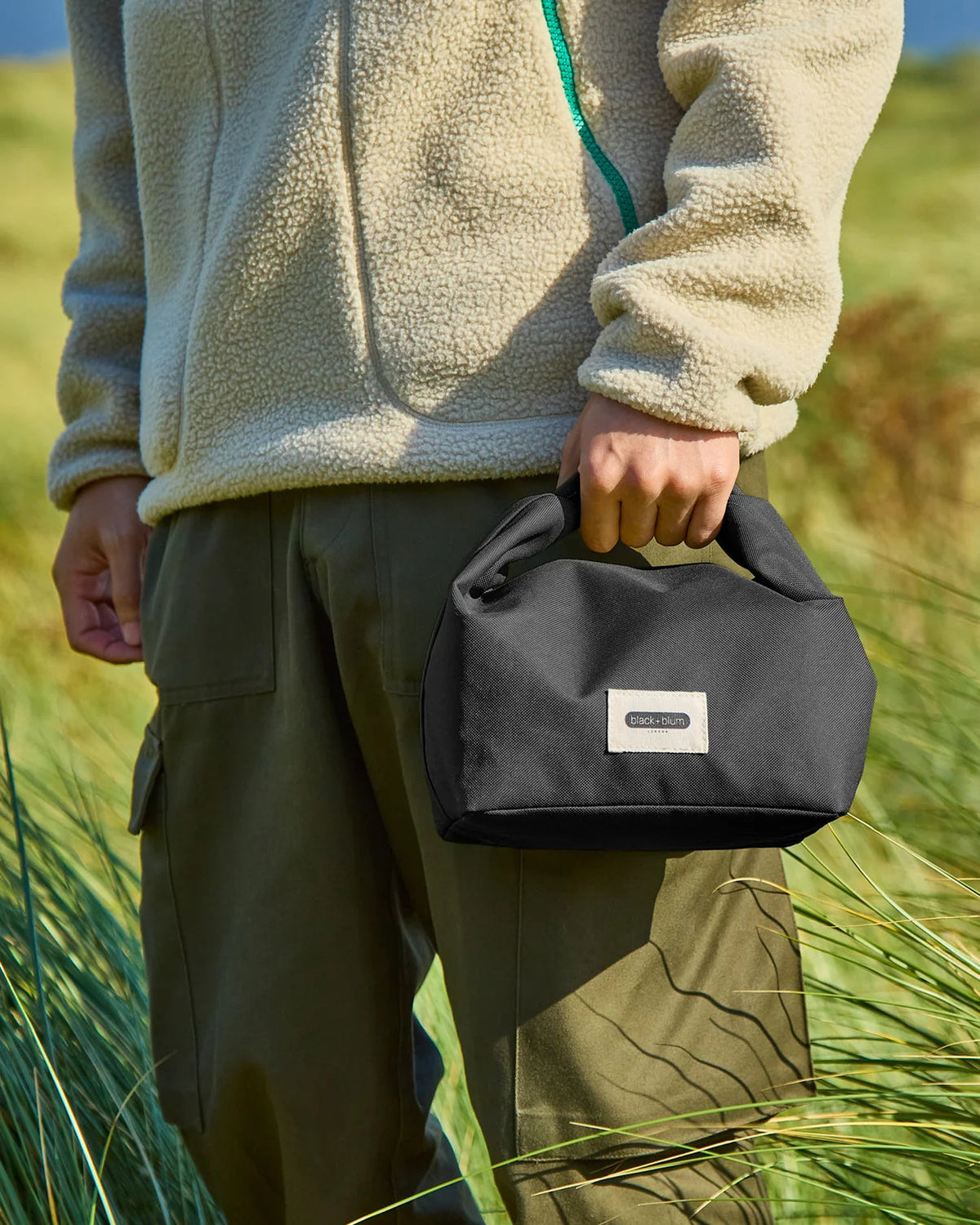 Maiden Cricket Black+Blum insulated lunch bag in black with roll-top closure held by a person in a grassy field