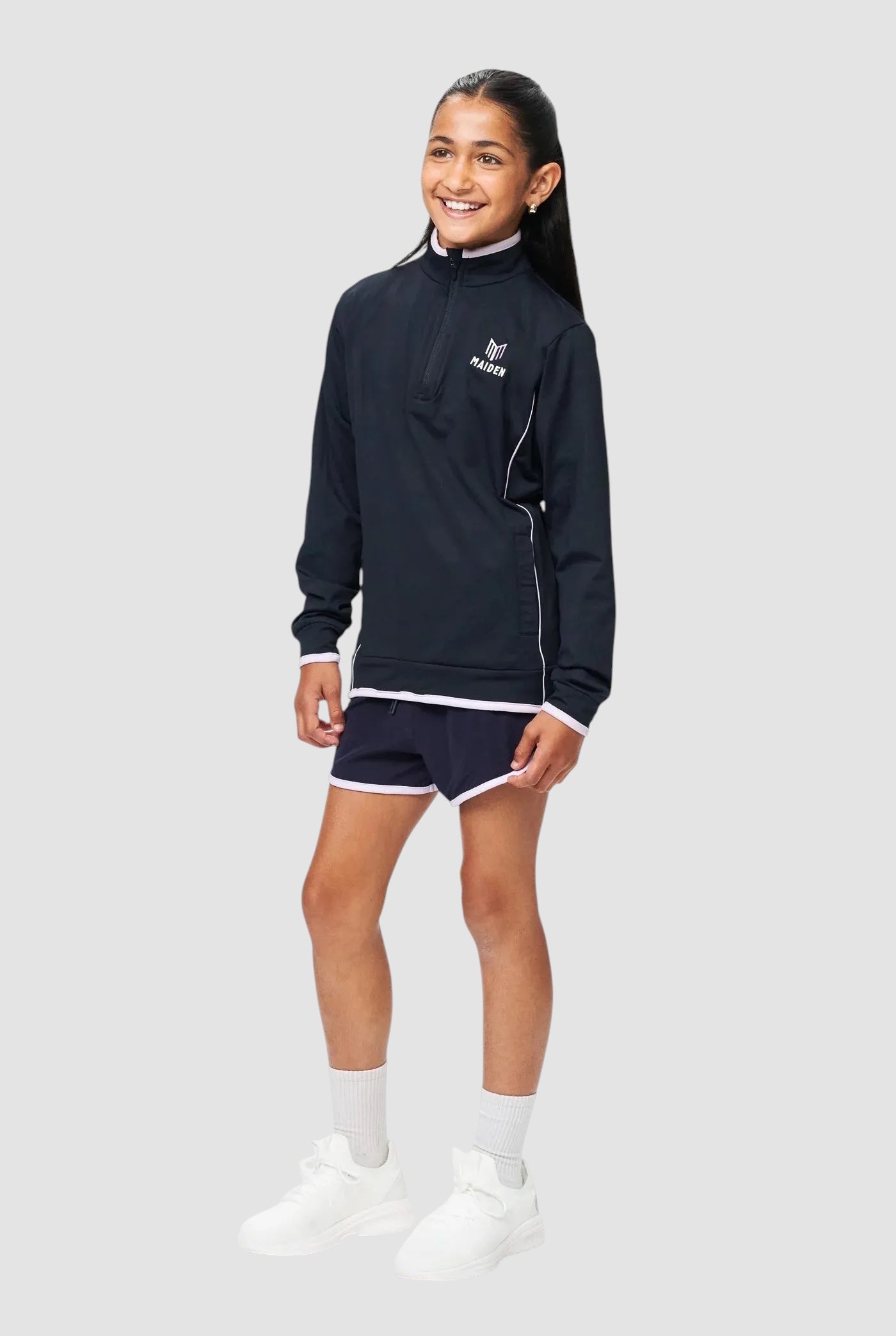 A young girl wearing a Maiden Cricket navy blue 1/4 zip midlayer with white trim and matching athletic shorts.