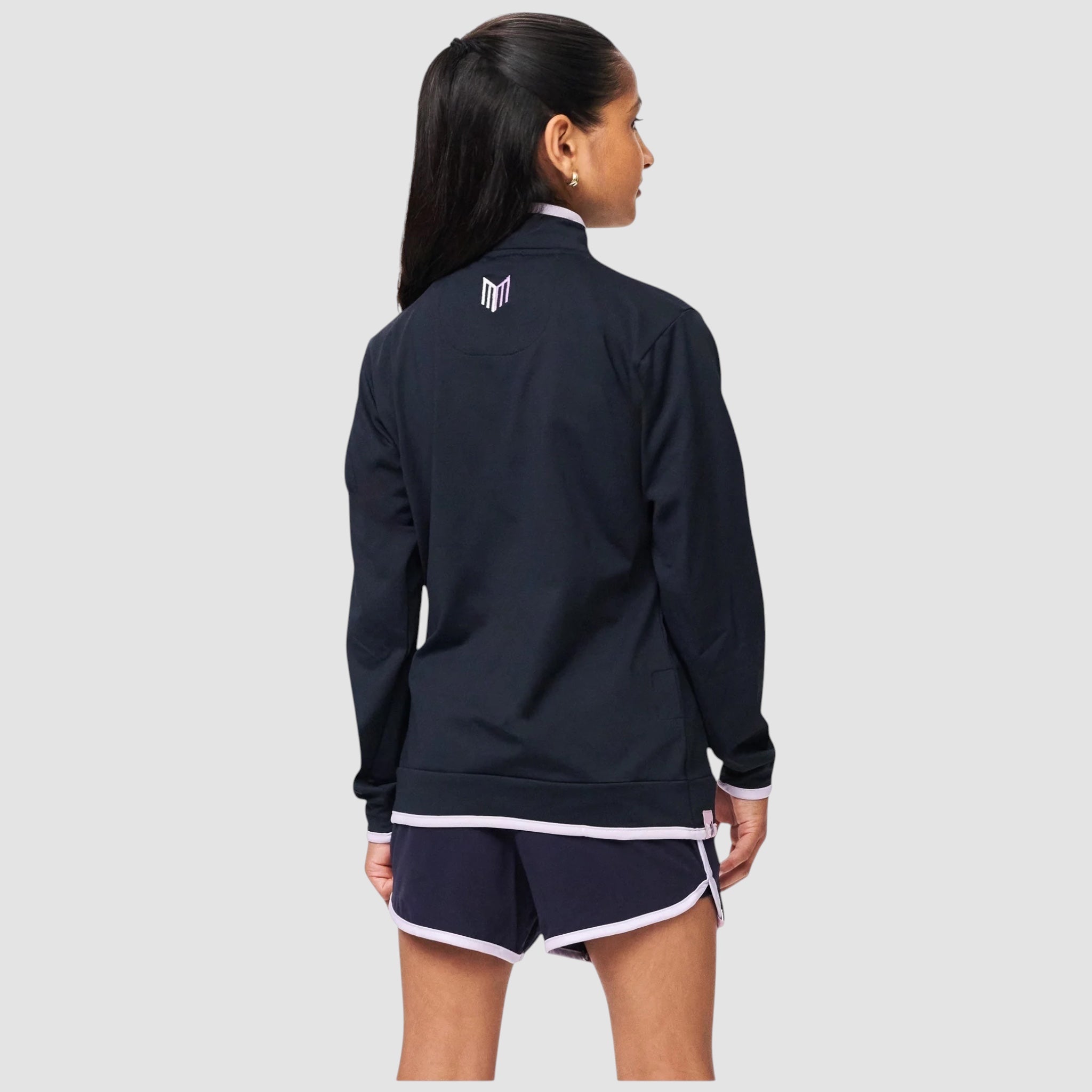 Back view of a girl wearing a Maiden Cricket navy blue quarter-zip midlayer with white trim and matching athletic shorts.