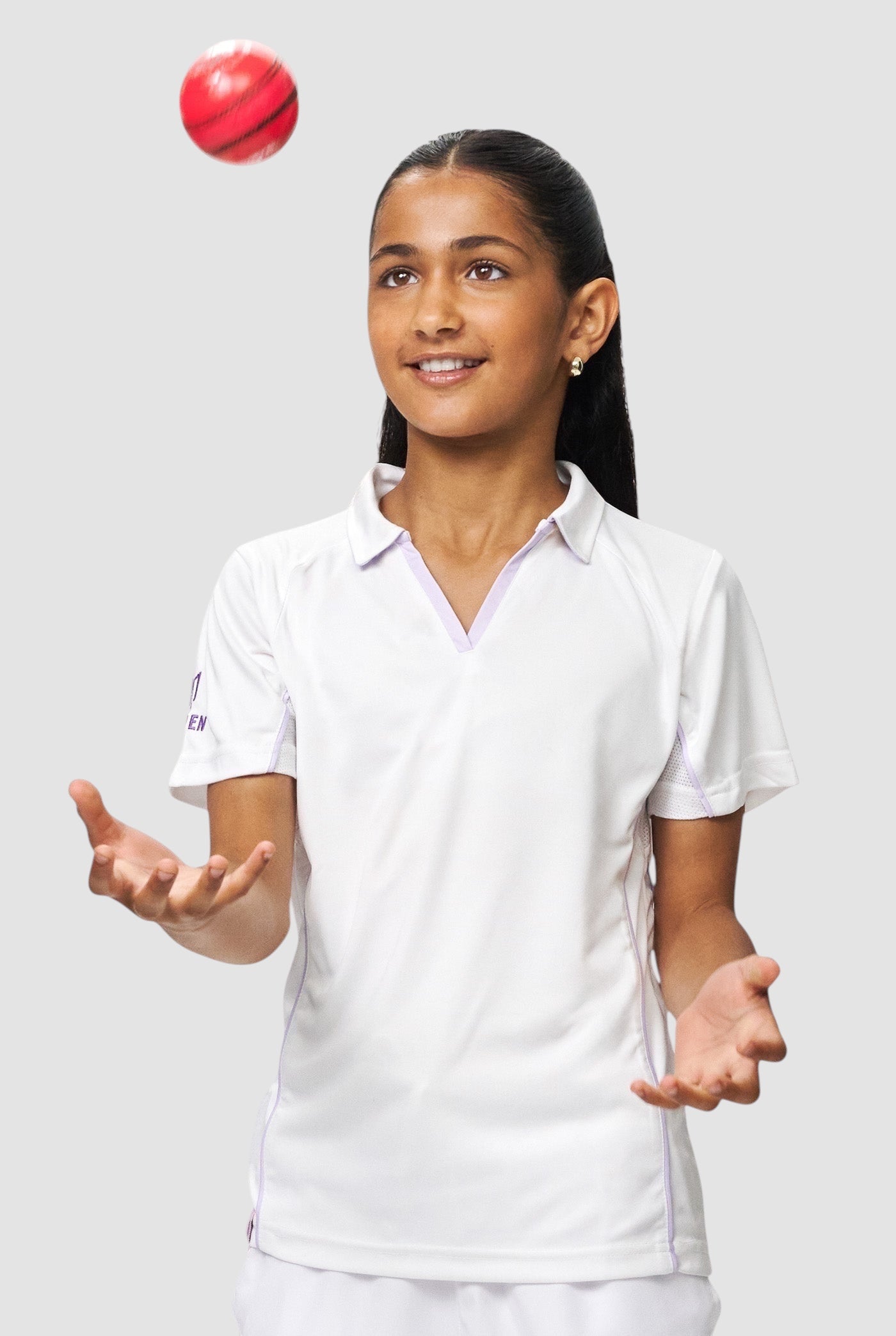 Young girl wearing a white Maiden Cricket performance polo with purple piping, tossing a red cricket ball in the air.