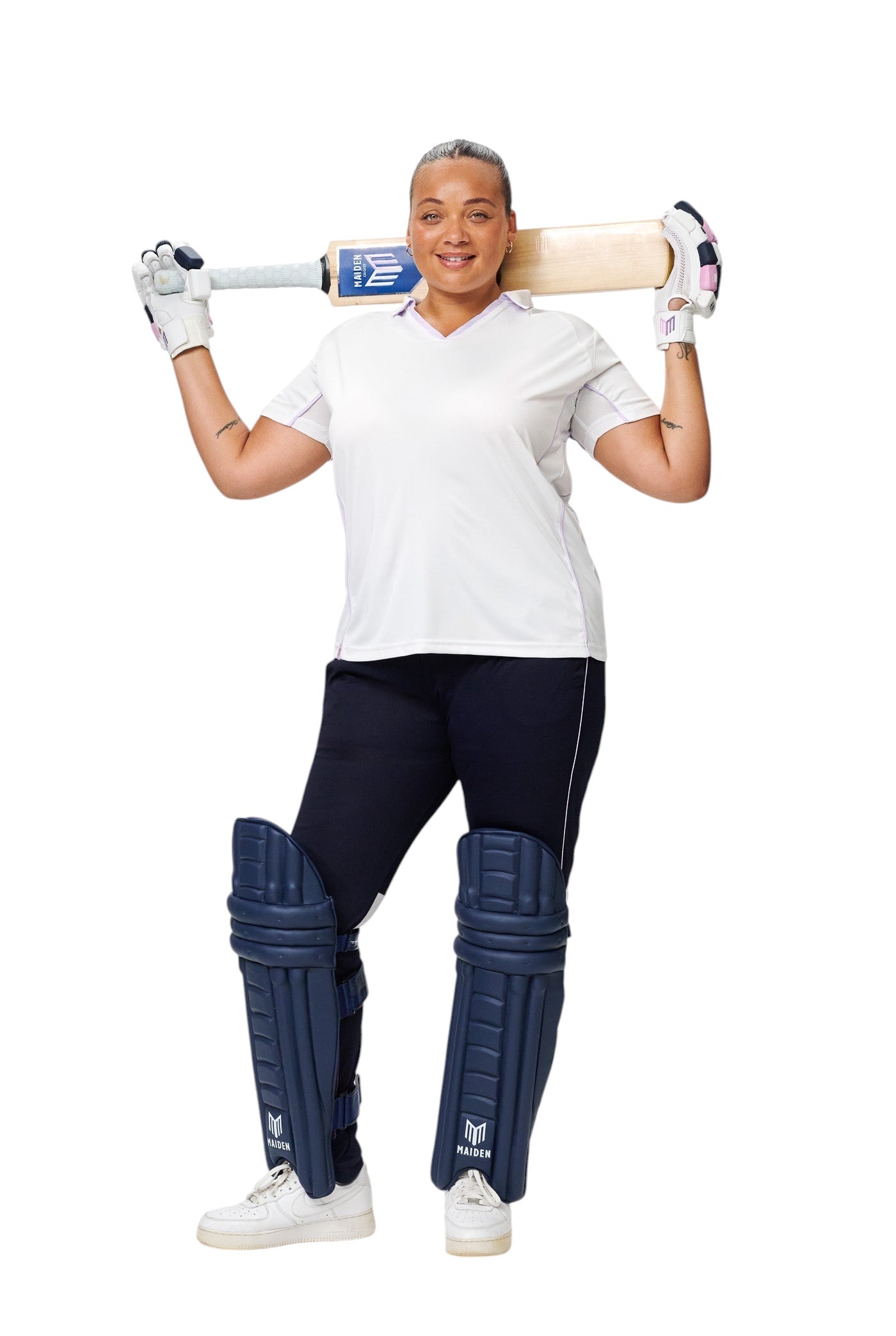 Female cricketer wearing Maiden Cricket navy batting pads, white shirt, and gloves while holding a cricket bat