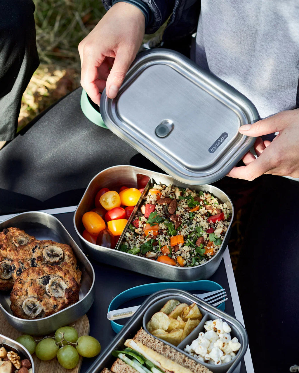 Maiden Cricket stainless steel lunch box with removable divider, filled with salad, fruit, and snacks outdoors.