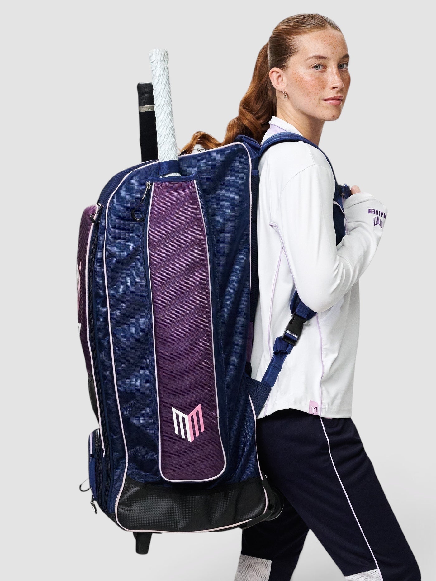 A female cricketer wearing a navy and purple Maiden Cricket kit bag with ergonomic straps and chest buckle.