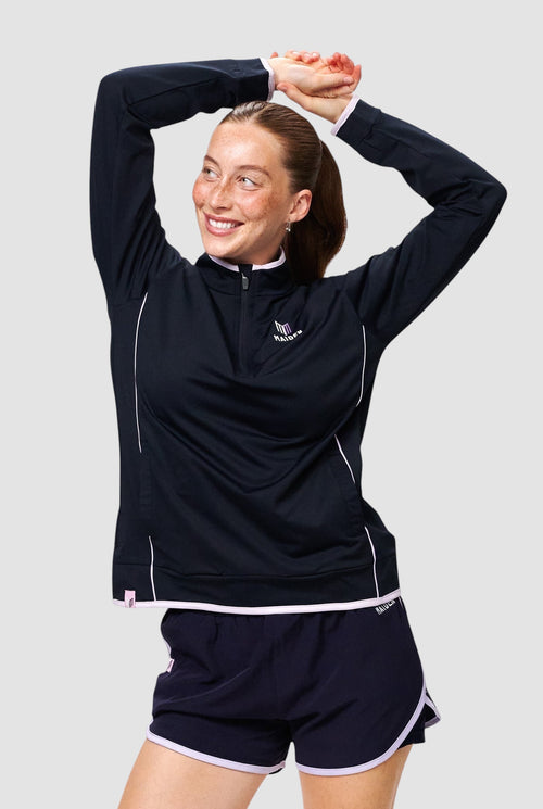 Maiden Cricket women's navy quarter-zip midlayer with light pink trim, worn by a smiling woman with hands overhead.