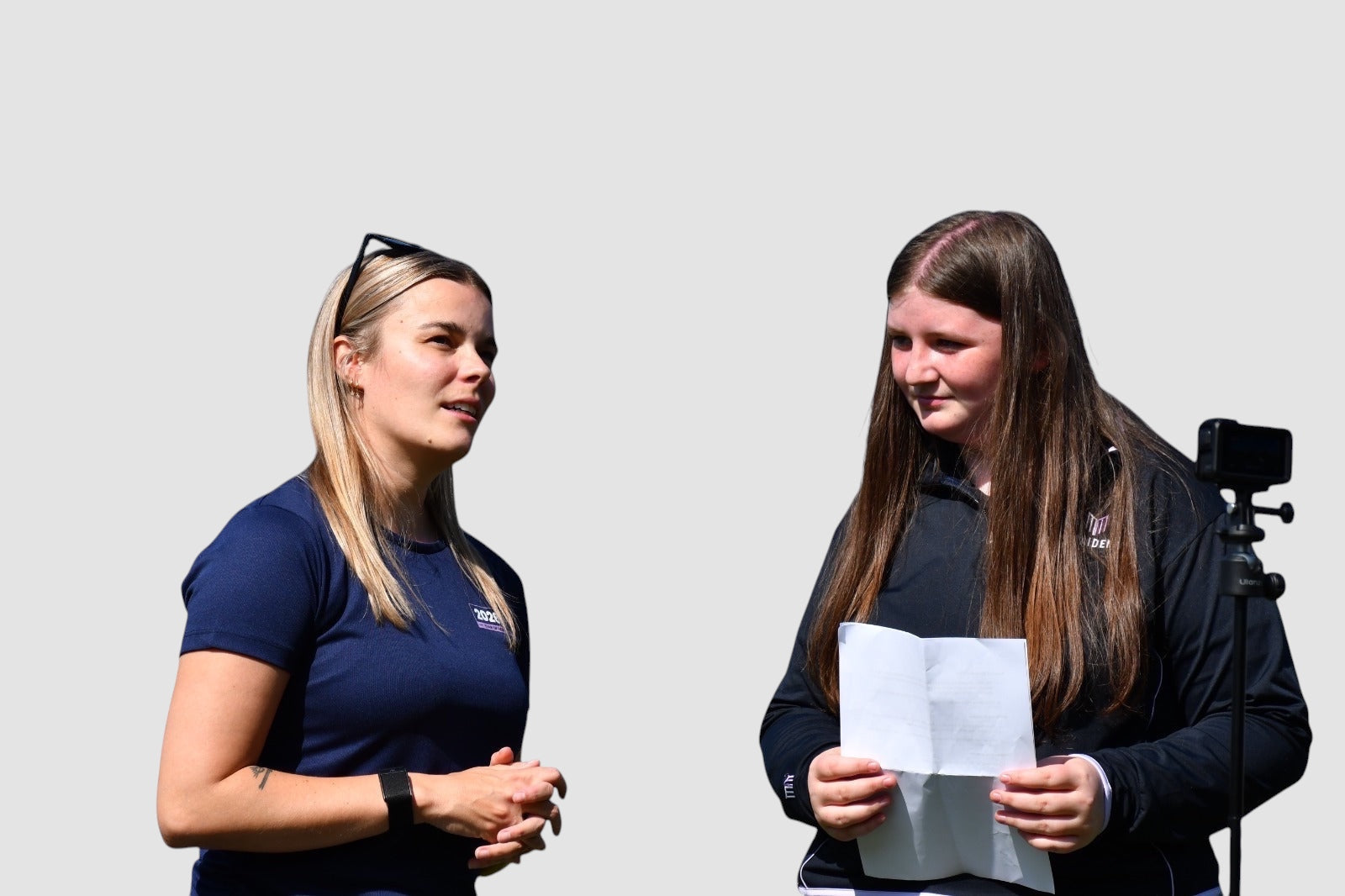Two women wearing Maiden Cricket branded sportswear, one in a navy blue t-shirt and one in a black long-sleeve top.