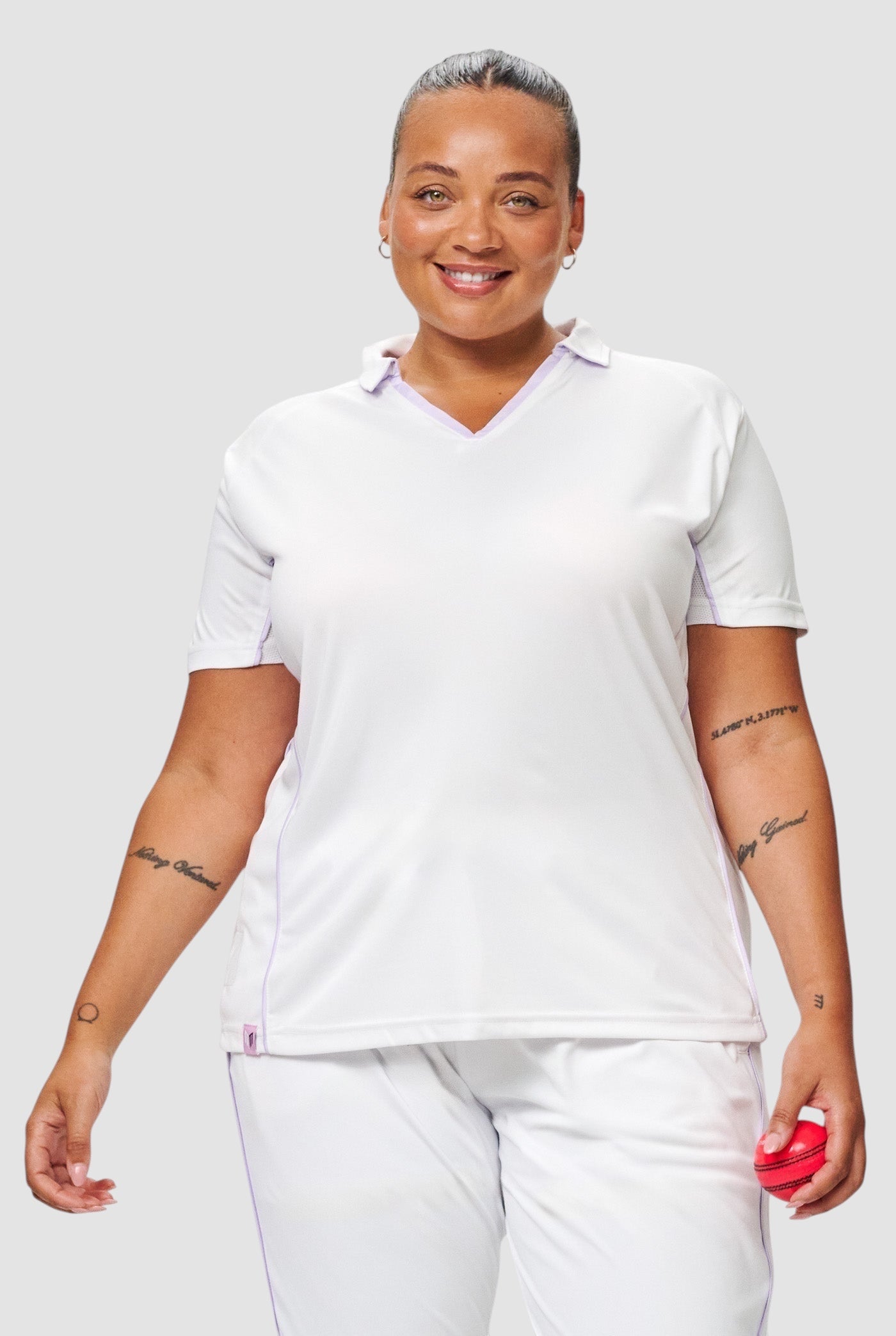 A smiling woman wearing a white Maiden Cricket Mid-On Top with a V-neck and purple trim, designed for female athletes.