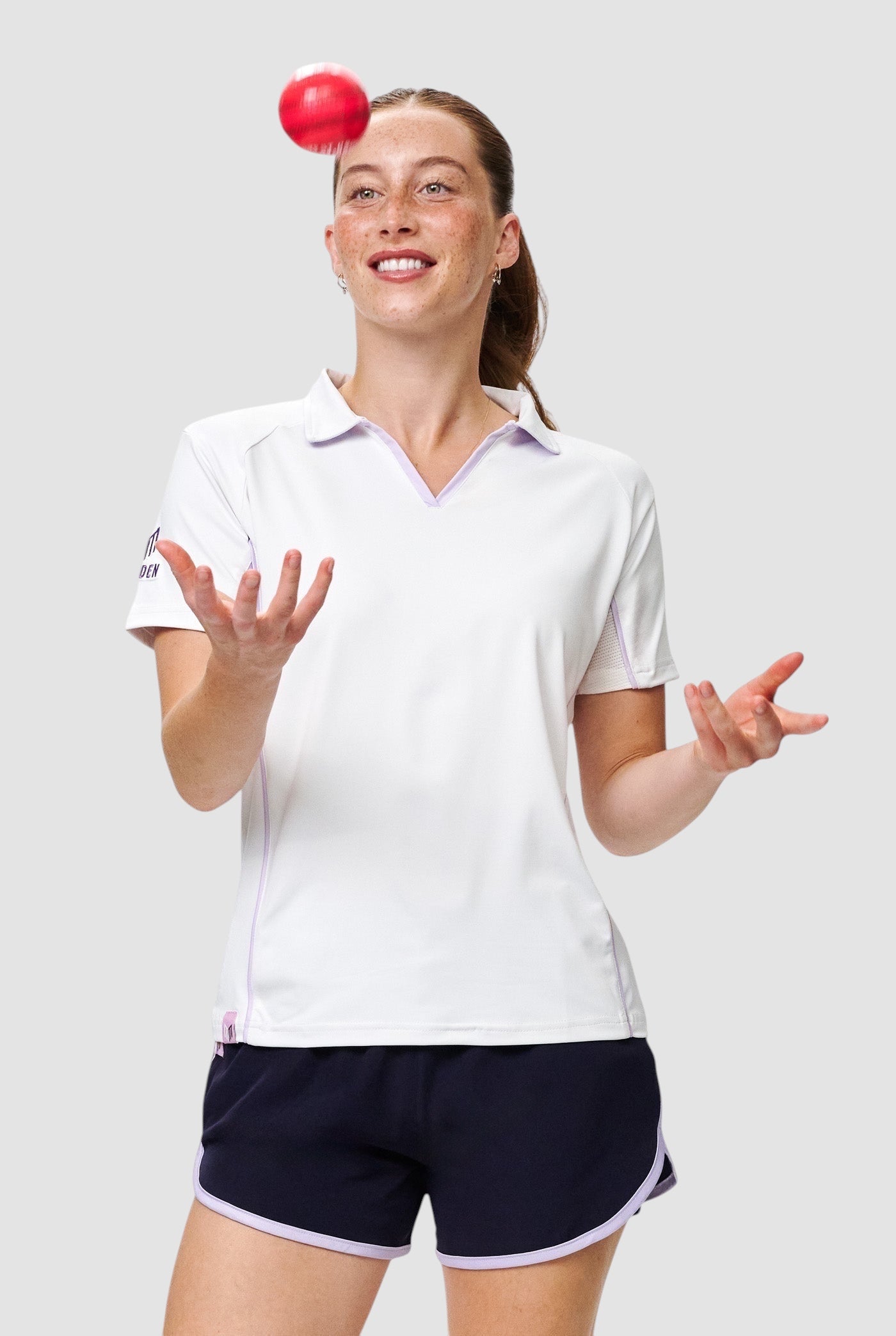 Maiden Cricket white V-neck performance top with mesh underarm panels worn by a female athlete juggling a red cricket ball