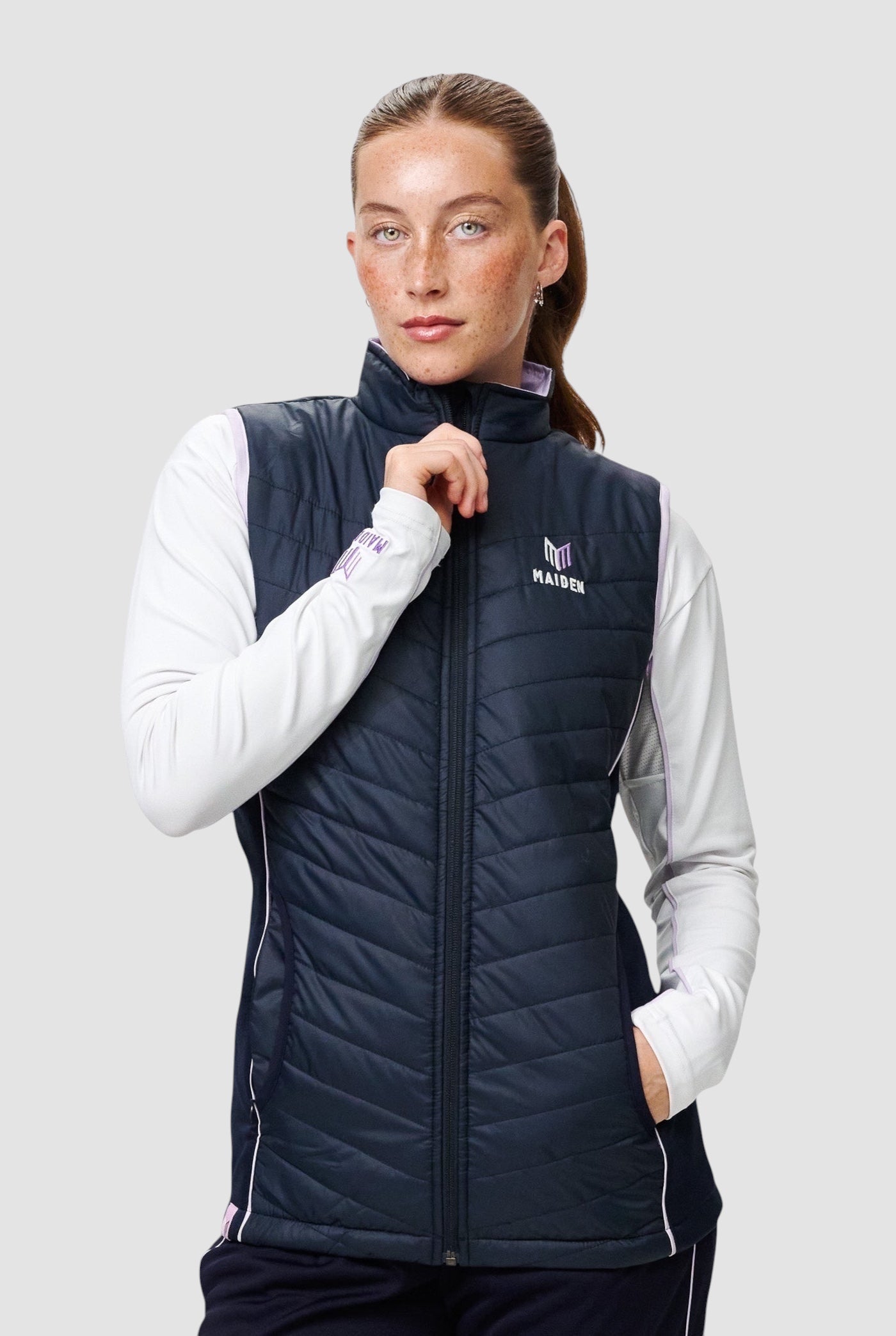 A woman wearing a navy blue Maiden Cricket Yorker Gilet with light purple accents over a white long-sleeve top.