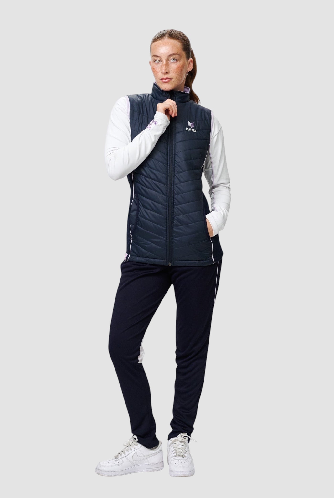 Woman wearing a navy Maiden Cricket Yorker Gilet with stretch side panels over a white long-sleeve top and navy pants